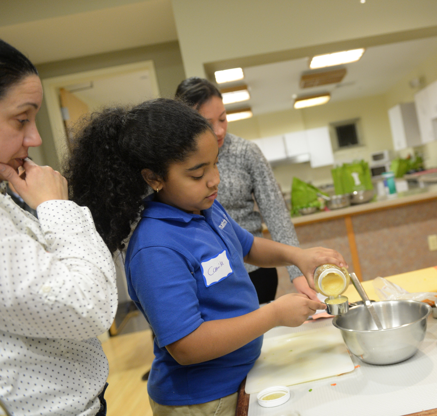 Girl measures ingredients for putting in bowl as two women watch