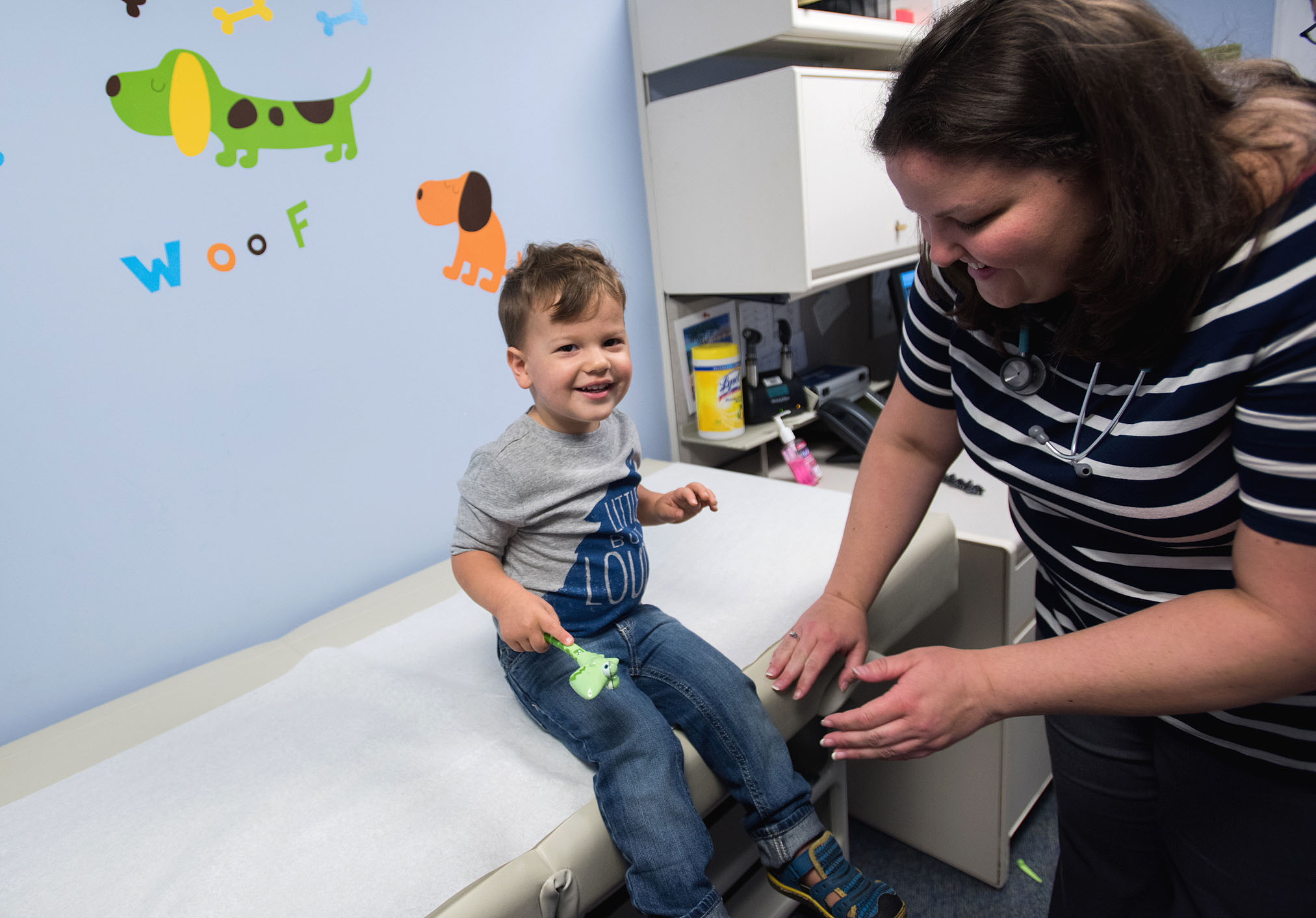 Toddler sitting on exam room table smiles as he is tended to by an adult woman
