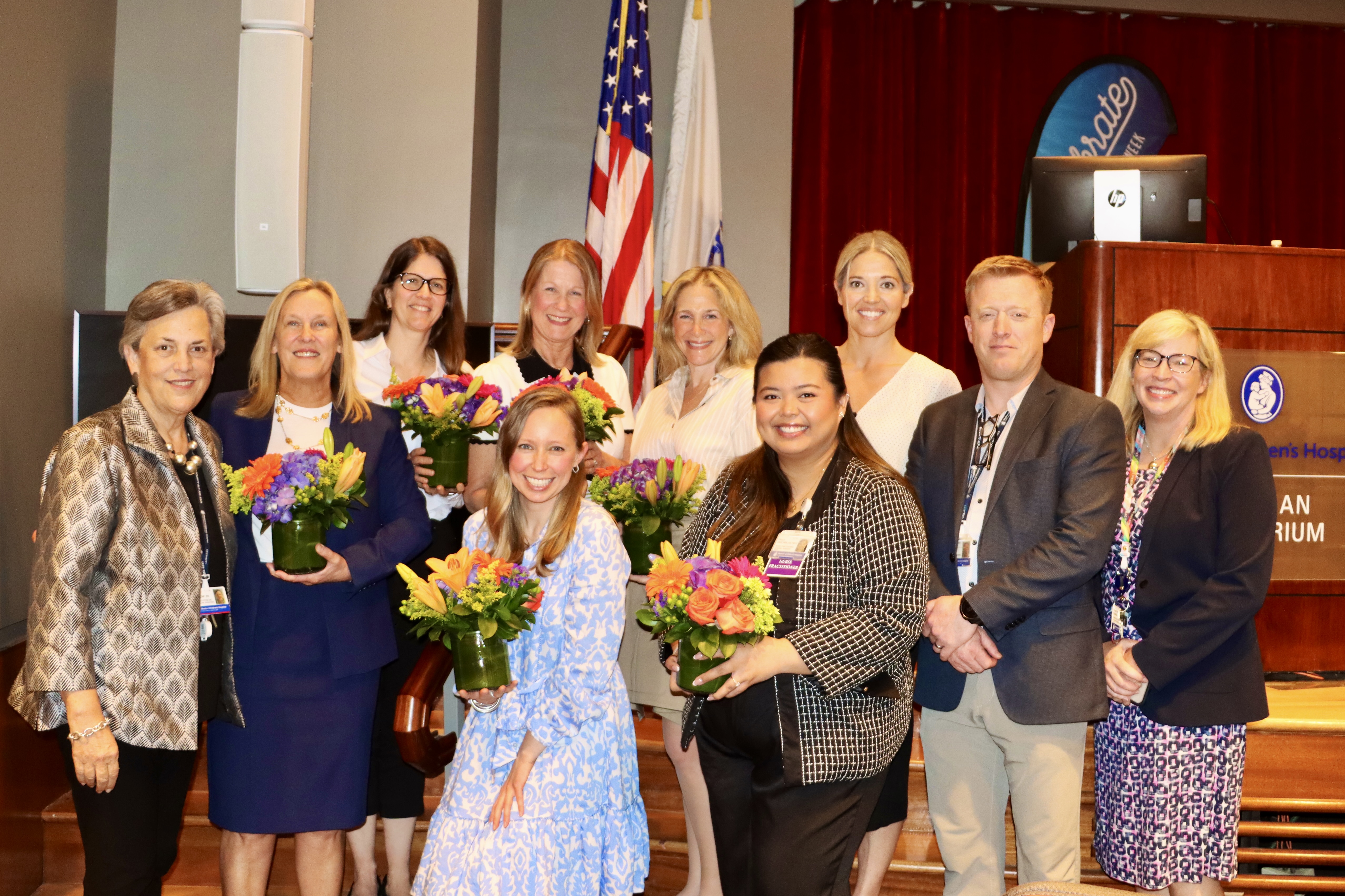 A group of nurses pose holding flowers on a stage in front of an american flag.