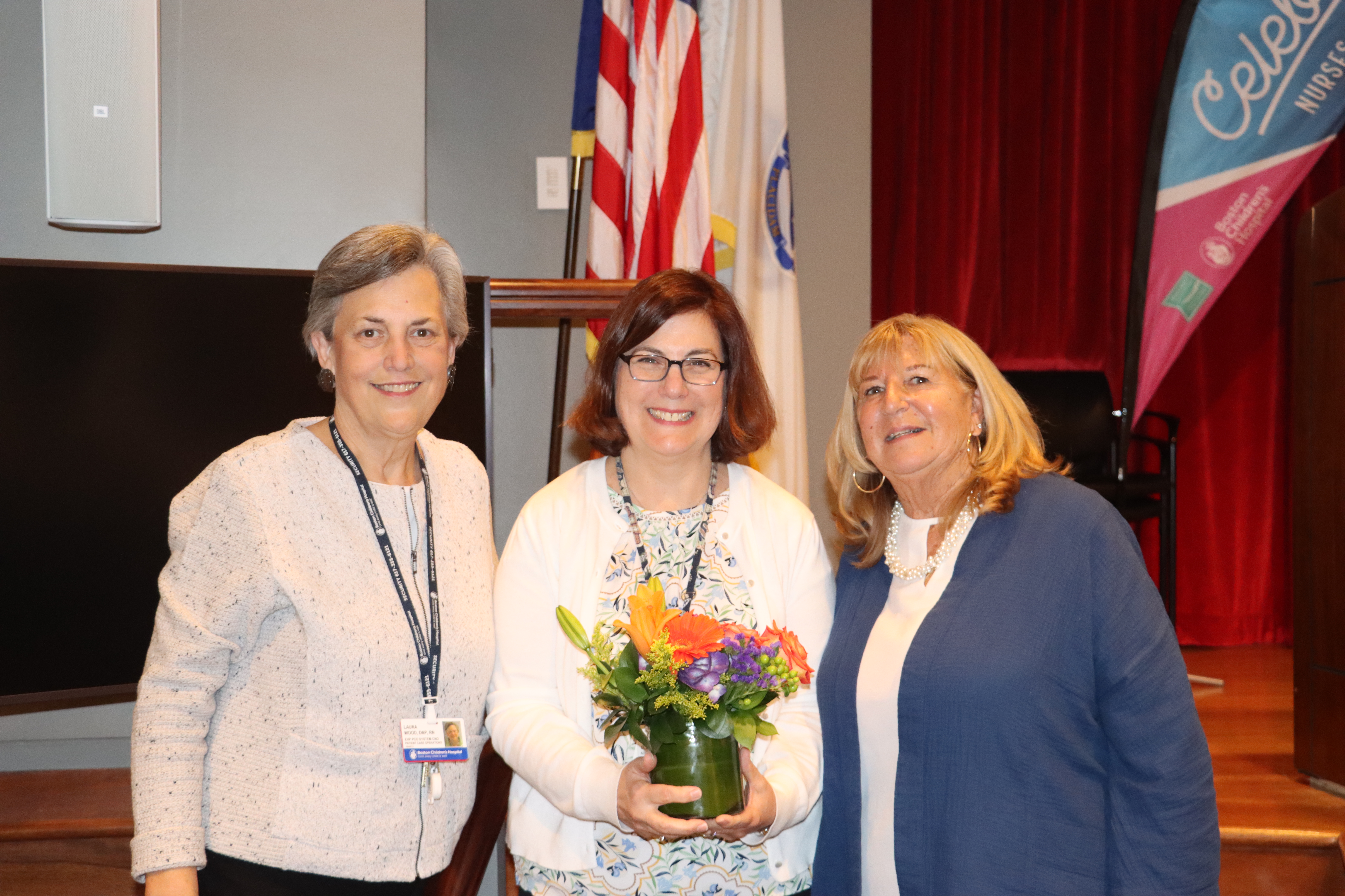 Three women, the middle one holding flowers, pose on a stage in front of an american flag.