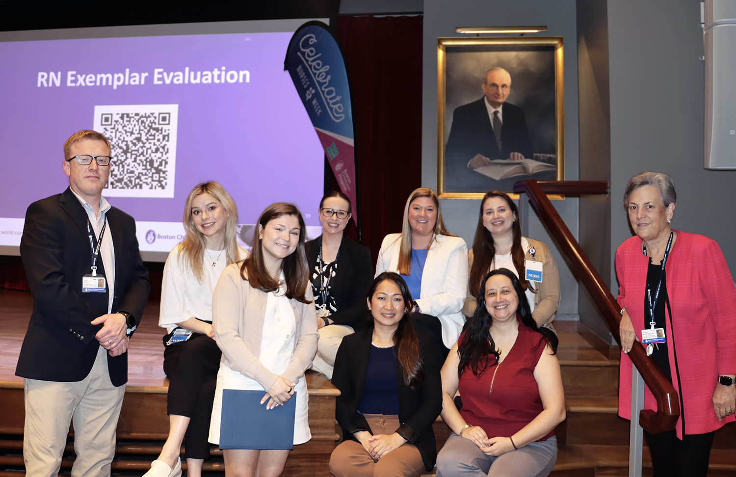 A group of RN exemplars pose together on a stage in front of a presentation slide with a QR code for RN examplar evalution and a painting of an old man in suit.