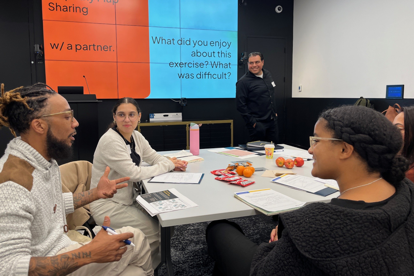 Youths sit around a table discussing an issue during seminar. Projector screen is at front of room.