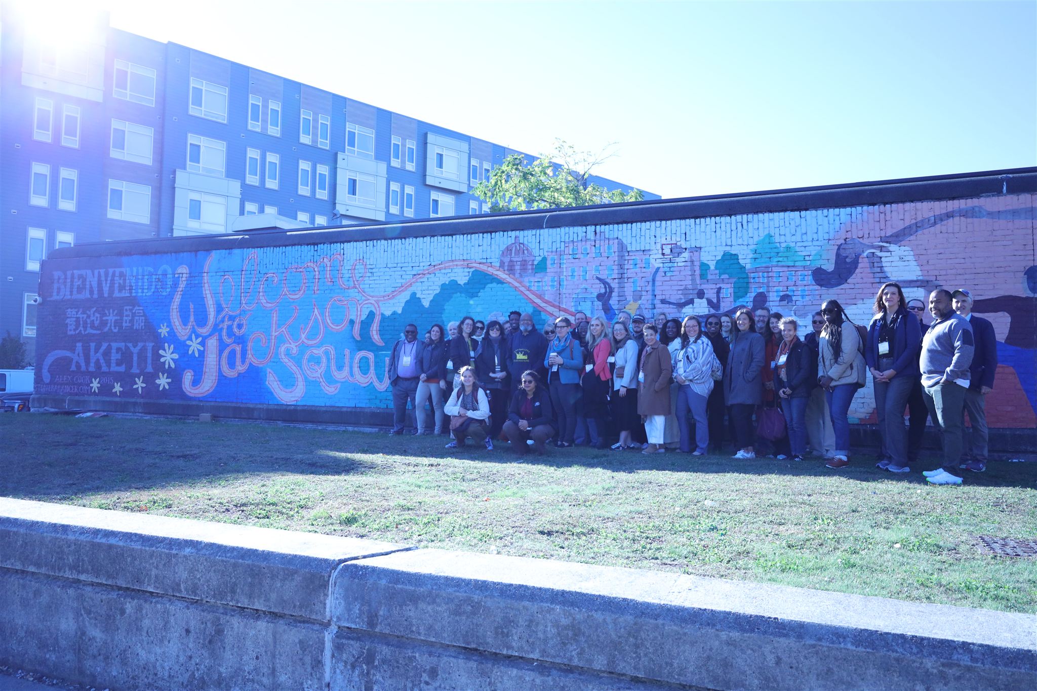 Large crowd stands in front of mural on side of the building as sun reflects off nearby window