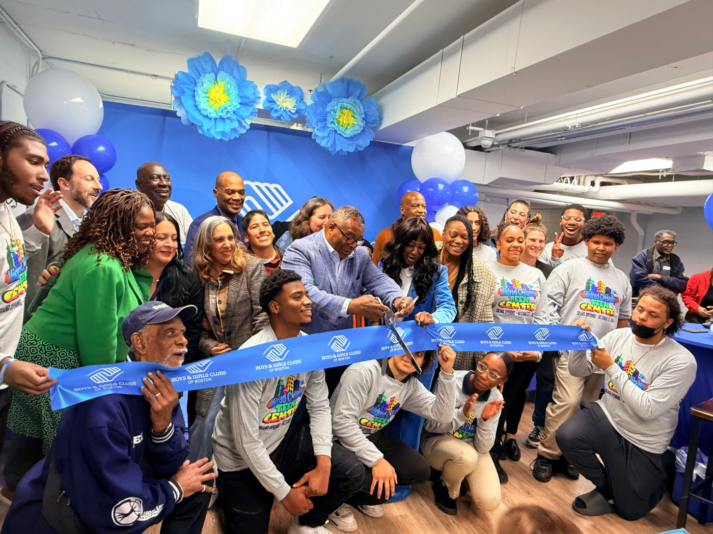 Man wearing glasses holds scissors to cut an oversized ribbon, with youth and adults standing behind him