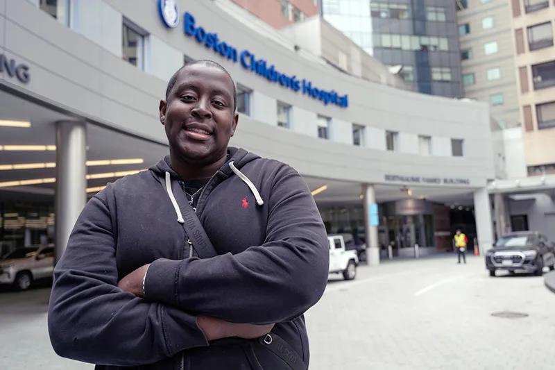 Young man, arms crossed, stands in front of main entrance to Boston Children's Hospital