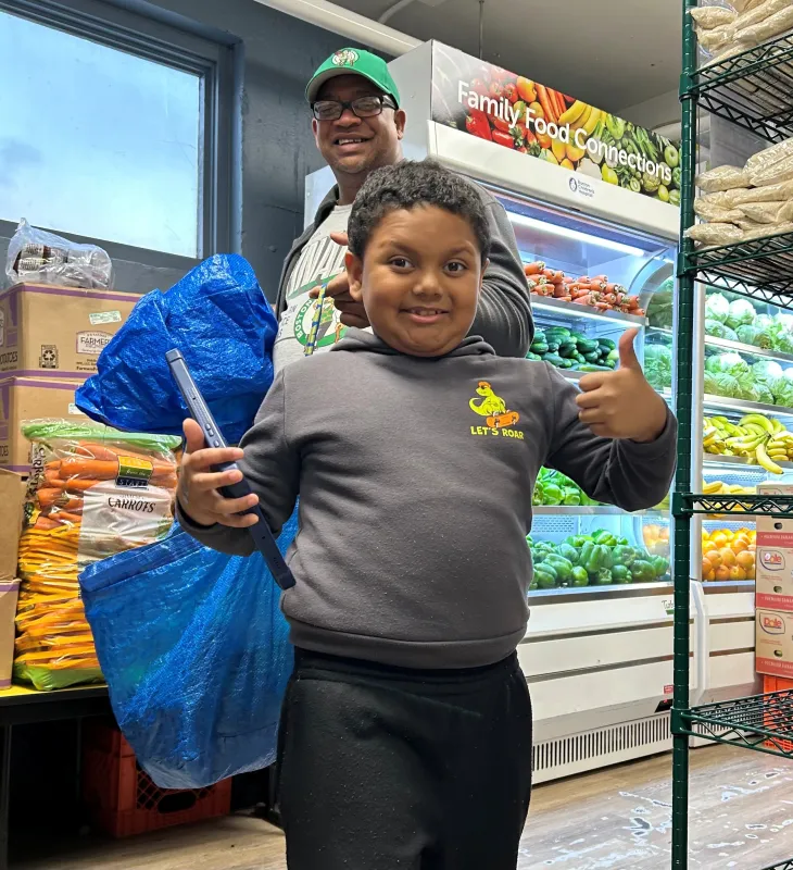 Younger boy gives thumbs-up sign when posing for photo. An older adult male stands behind him, carrying bags of groceries.