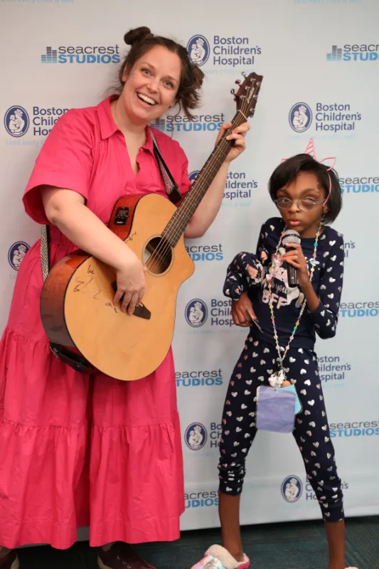 Smiling woman holding guitar stands on left, next to young girl holding a microphone to her mouth