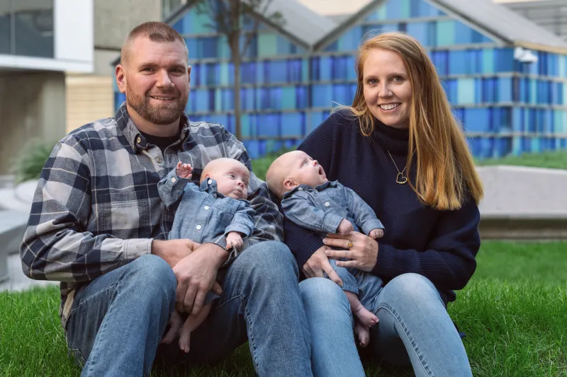 Man with short hair and woman with long hair sit on grass, each holding one baby