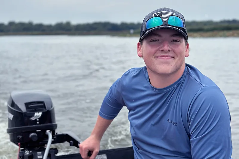 Boy wearing blue shirt, baseball hat and sunglasses poses on a boat on a body of water
