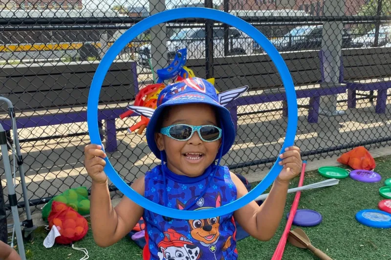 Child wearing sunglasses and helmet holds up a hula hoop.