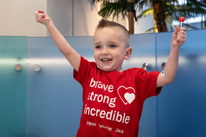 Boy wearing T-shirt holds his arms up, has lollipop in his left hand